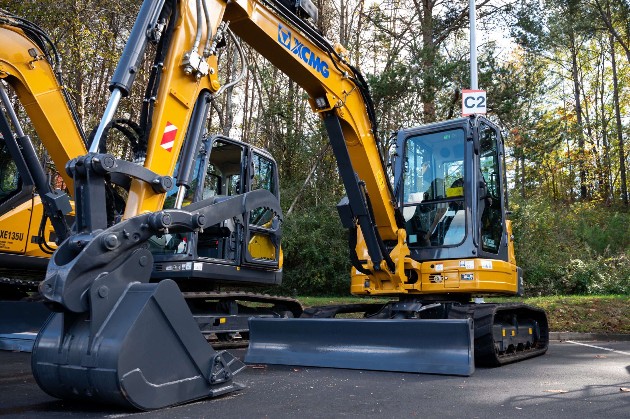 Two yellow excavators with cabs, labeled XCMG, are parked on a paved surface near a wooded area. The excavators have large buckets and digging arms positioned in the foreground.
