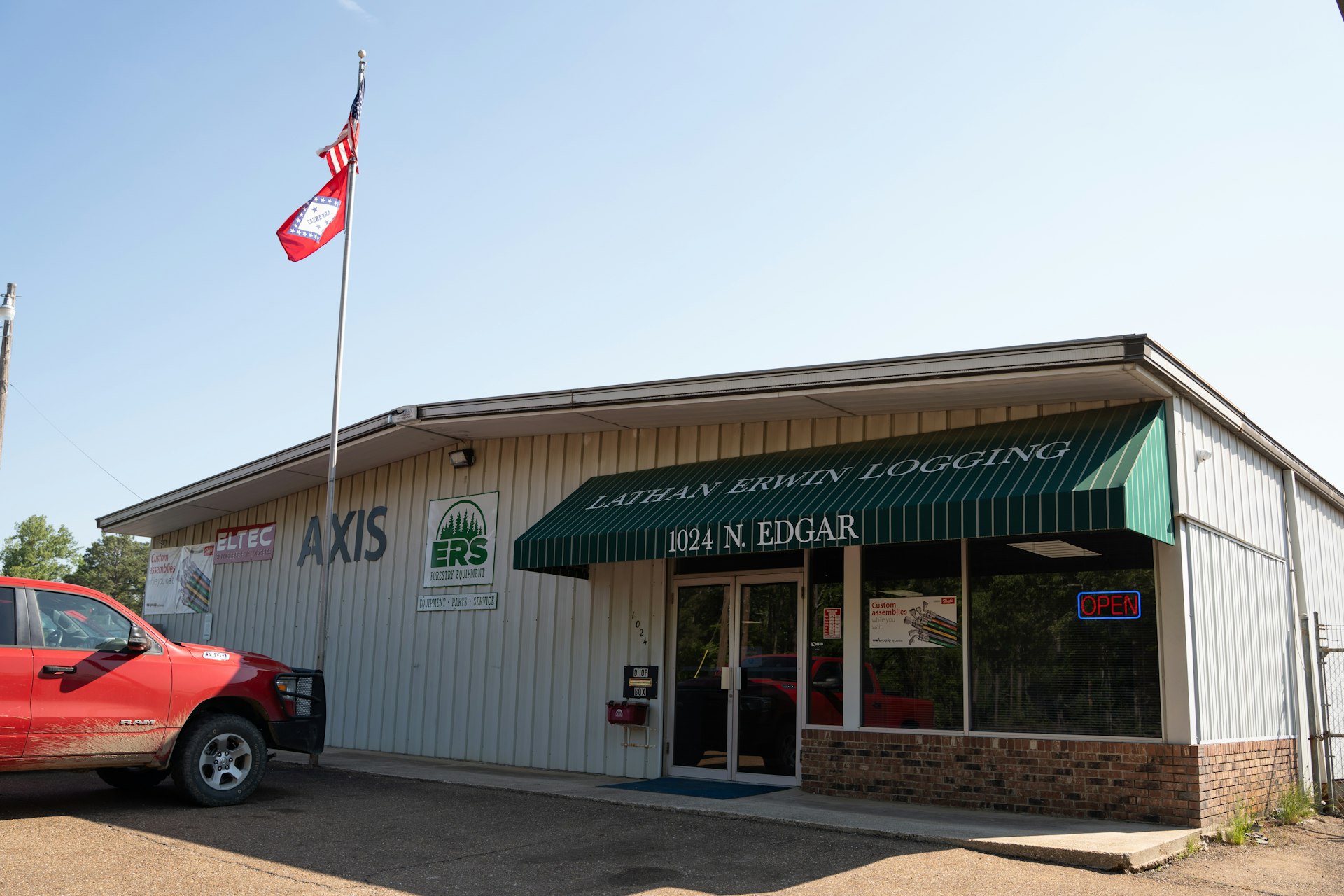 A small commercial building with a green awning reading Lathan Erwin Logging 1024 N. Edgar. A red truck is parked in front, and two flags fly on a pole. The building has signs for AXIS and ERS.