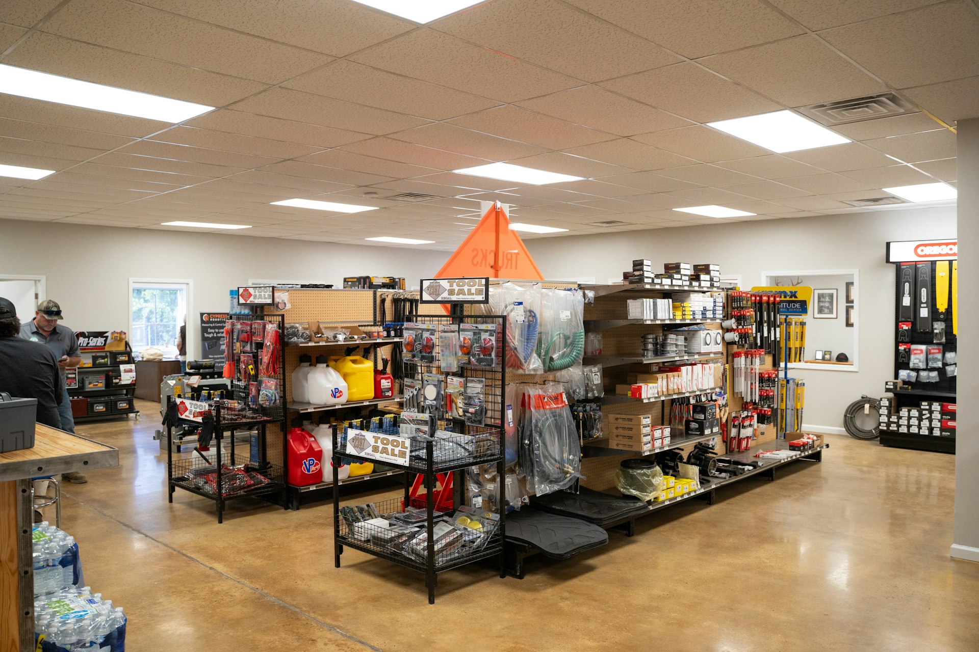 A hardware store interior with shelves displaying tools, equipment, and accessories. The floor is polished brown, and fluorescent lights illuminate the space. A person is at the counter on the left.