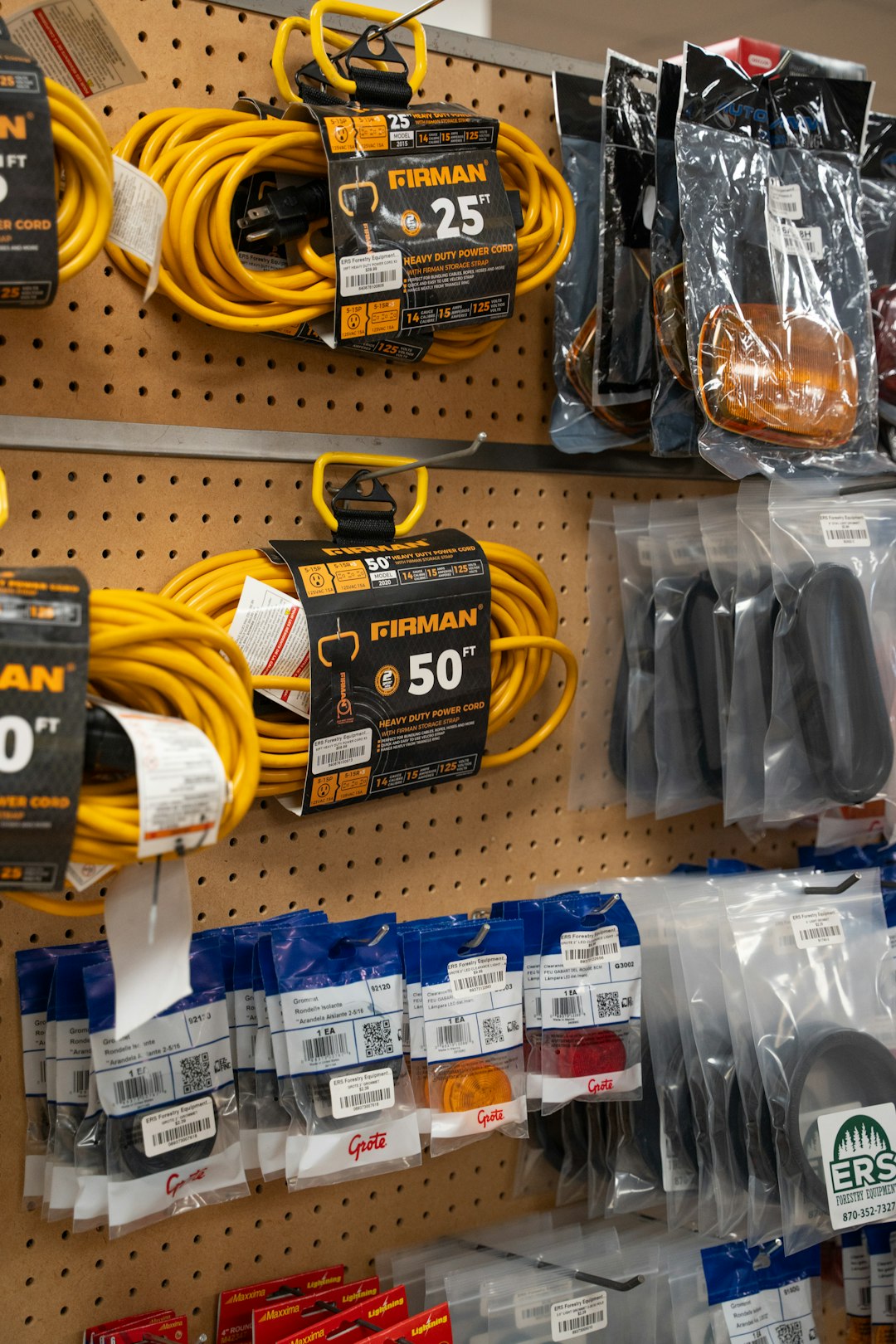 Yellow extension cords and various packaged hardware items hang on a pegboard display in a store. The cords are labeled FIRMAN and come in 25- and 50-foot lengths. Other items are in clear or blue plastic packaging.