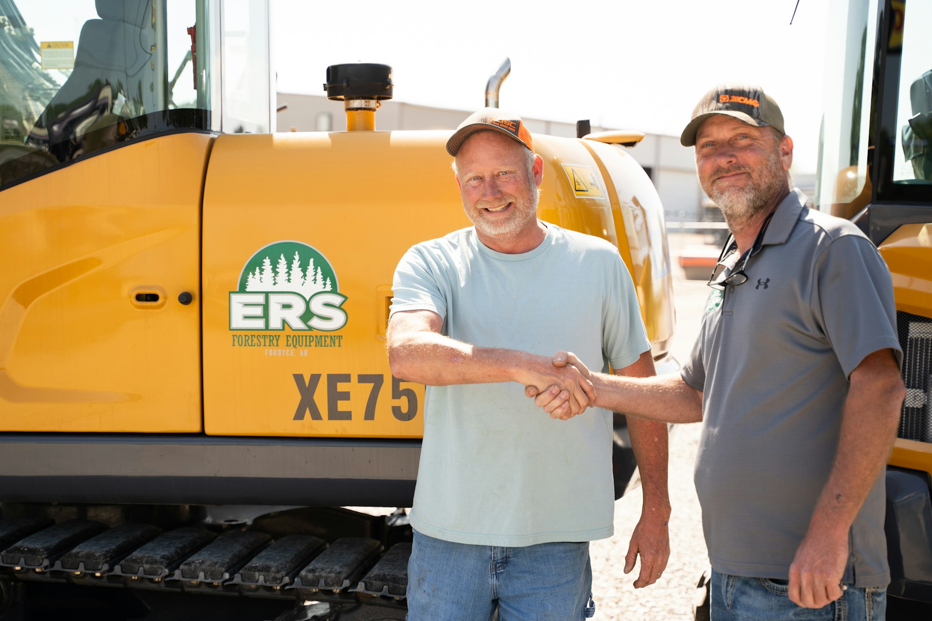 Two men in casual clothes shake hands and smile in front of a yellow construction vehicle labeled ERS Forestry Equipment XE75 on a sunny day.