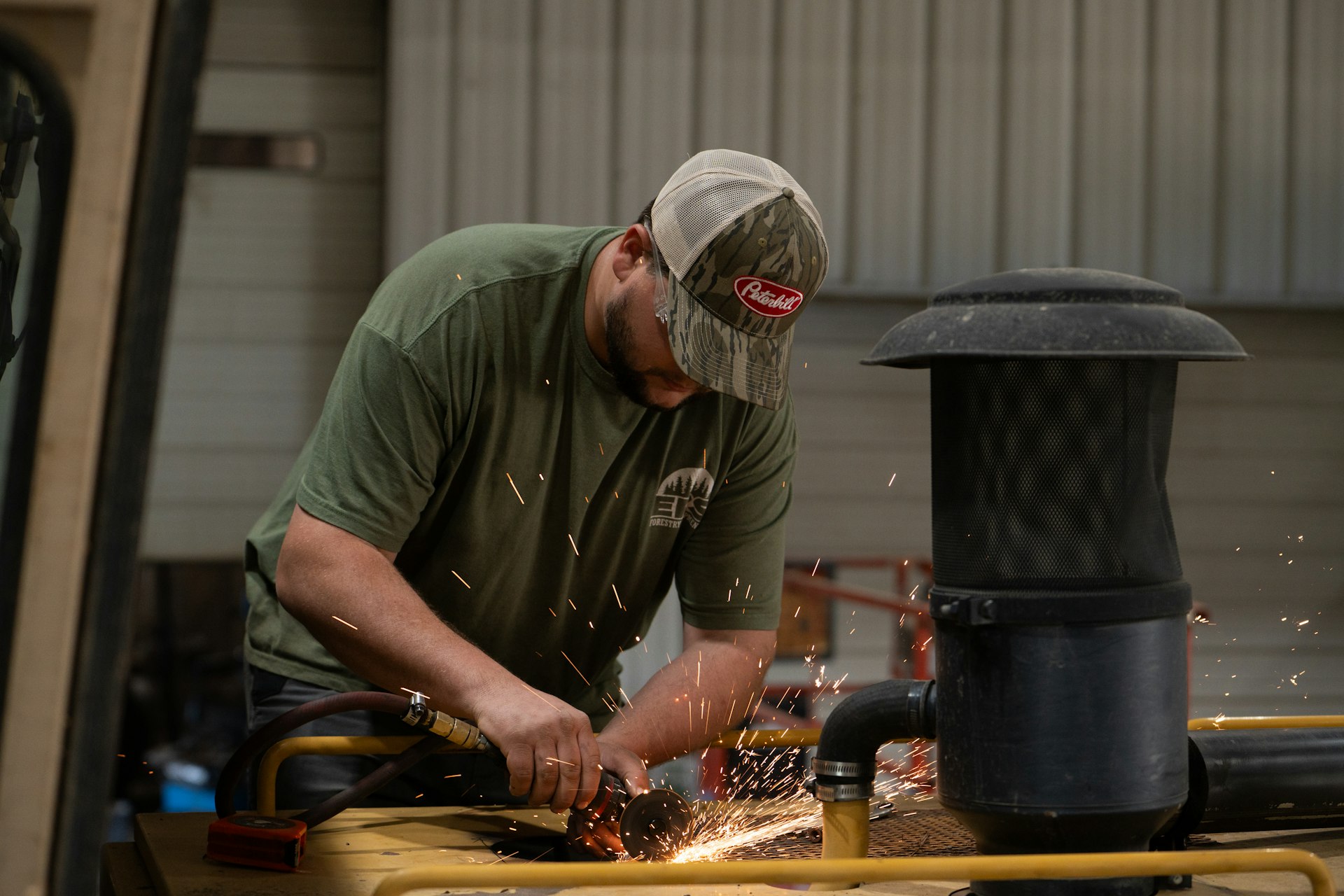 A man wearing a green t-shirt and beige cap uses a power tool to grind metal, producing sparks, in an industrial workshop setting.