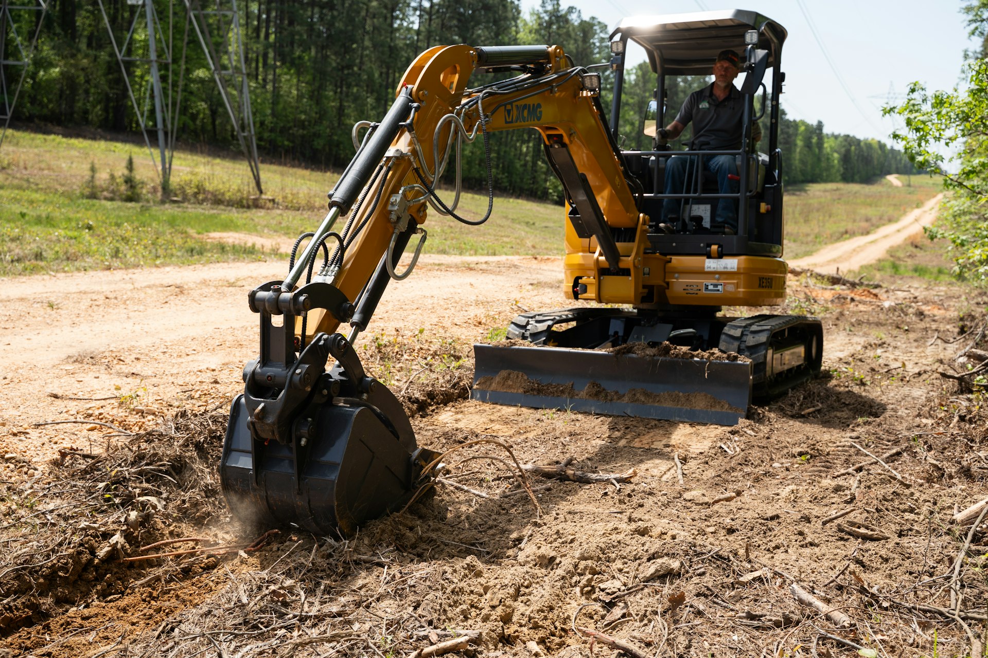 A man operates a small yellow excavator, digging into dry, rough soil beside a dirt road surrounded by trees and greenery.